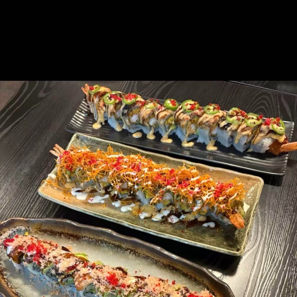 Three long plates of sushi rolls, each with unique toppings and sauces, on a dark wooden table.
