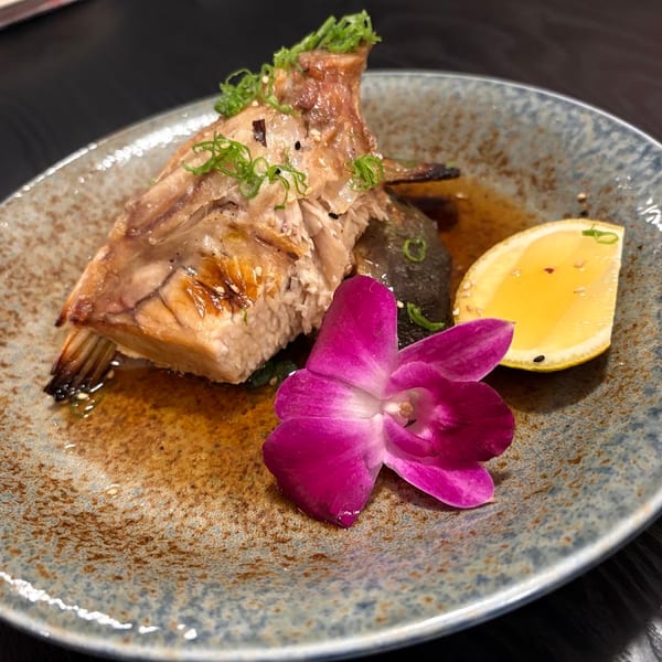 Grilled fish with green onions, lemon, and a purple orchid on a speckled plate.